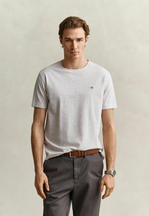 Jeune homme aux cheveux courts et bruns porte un t-shirt gris clair, un pantalon foncé, une ceinture marron et une montre-bracelet, debout devant un fond uni.