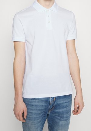 Mężczyzna w gładkiej, białej koszulce polo z krótkim rękawem i trzema guzikami, w zestawieniu z niebieskimi dżinsami, stojący na białym tle.