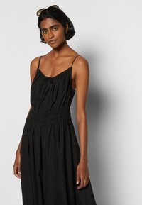 Vestido preto sem mangas com corpete franzido, alças ajustáveis e cintura elástica. O tecido parece leve e fluido.