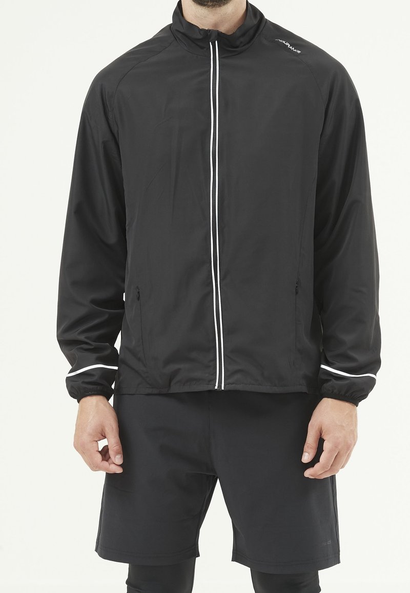 Homme portant une veste noire à fermeture éclair avec bordure blanche et un short noir sur un fond uni.