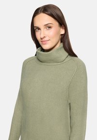 Pull à col roulé vert à la texture douce, avec une coupe ample et des détails côtelés au niveau du col et des épaules.
