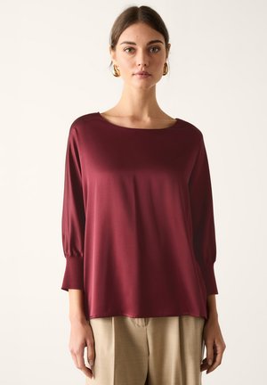 Ung kvinde iført en løs bordeaux satinbluse med trekvartærmer og beige bukser, der står mod en ensfarvet baggrund.