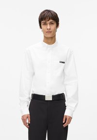 Balts pogcauruma krekls ar kabatu un melnu logotipa uzlīmi, apvienots ar melnu jostu un tumšiem biksēm. Gluda tekstūra, pielāgota piegriezuma.