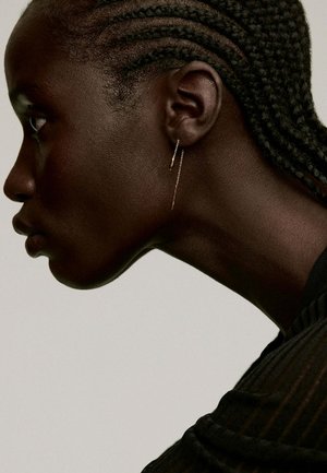Un pendiente de gota dorada cuelga de un peinado con el cabello corto, con un tono de piel liso y oscuro y una vista de perfil que resalta una mandíbula bien definida.