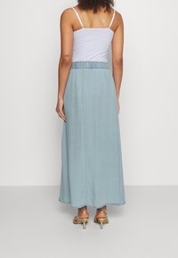 Kvinna i ljusblå högmidjad maxikjol, vit tank top och guldiga sandaler med klack, som står mot en enkel bakgrund.