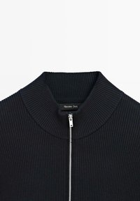 Maglione a coste nero con colletto alto e zip frontale argentata, etichetta del marchio visibile all'interno del colletto.