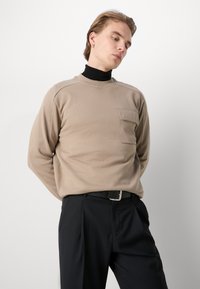 Camicia beige a maniche lunghe con taschino sul petto, texture liscia, scollatura rotonda e maniche raglan, indossata sopra un dolcevita nero, insieme a pantaloni neri.