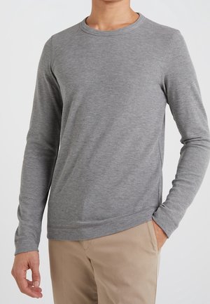 Chemise grise à manches longues avec une texture côtelée, col rond et poignets ajustés, associée à un pantalon beige clair.