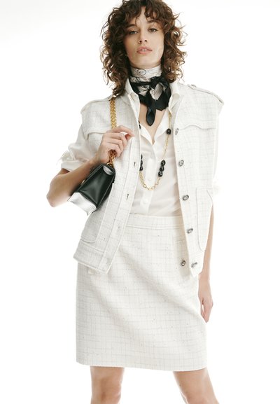 Vrouw met een witte, gestructureerde vest en rok, een blouse met opgerolde mouwen, een zwarte sjaal met patroon, een gouden ketting en een zwarte handtas.