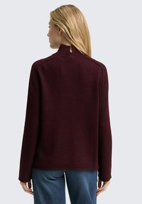 Bordeaux gebreide trui met een hoge hals, raglanmouwen en geribde manchetten. Zachte textuur, gecombineerd met blauwe denimbroek.