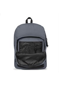 Mochila gris con un bolsillo frontal con cremallera, que cuenta con organizadores de malla y un diseño redondeado. El material es sintético con una textura mate.