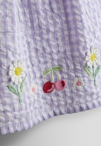 Tissu vichy violet clair avec des marguerites brodées et une paire de cerises rouges le long du bord.