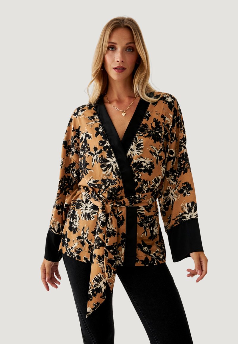 Blusa envolvente floral en un tono marrón cálido con patrón floral negro. Presenta un profundo escote en V, mangas largas y un cinturón atado en la cintura. Textura de tela suave.