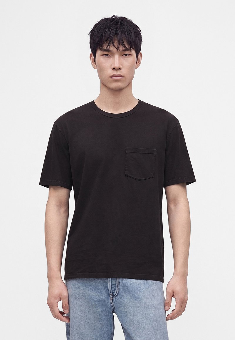 Jeune homme aux cheveux noirs courts portant un t-shirt noir uni avec une poche poitrine et un jean bleu clair, debout devant un fond blanc.