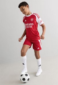 Rode voetbalshirt met witte mouwen en accenten, voorzien van een gestructureerd patroon. Rode shorts. Speler draagt witte sokken en staat op een zwart-witte voetbal.