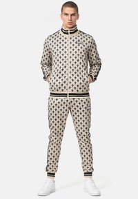 Beige tracksuit with a floral pattern, featuring a zip-up jacket and elastic waistband pants. Black accents on cuffs and collar. White sneakers.