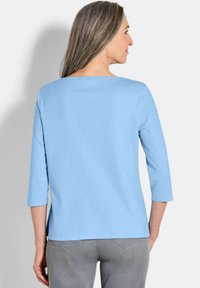 Himmelblaues Oberteil mit 3/4-Ärmeln aus weichem Material, Rundhalsausschnitt und lässiger Passform, kombiniert mit grauen Hosen.