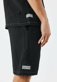 Pantalones cortos negros con textura y patrón de rayas horizontales, con costuras blancas y etiquetas de marca en el costado y el dobladillo.