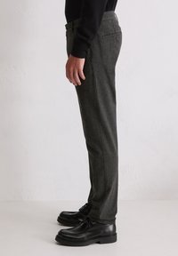 Mann steht in grauen karierten Hosen, schwarzen Leder-Schnürschuhen und einem schwarzen Langarmoberteil vor einem schlichten weißen Hintergrund.