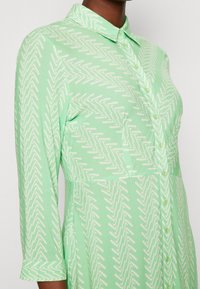 Robe chemise vert menthe avec boutonnage sur le devant, col et manches trois-quarts. Présente un motif en zigzag blanc sur toute la surface et une coupe légèrement évasée.