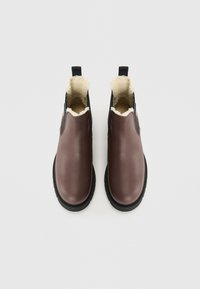 Et par brune lær slip-on støvler med sorte elastiske sider, sorte såler og krämfleece-fôr, vist fra oven på hvid baggrund.