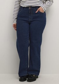 Jeans anchos de color azul oscuro con una cintura alta, bolsillos delanteros y costuras en contraste; combinados con una camisa de rayas de manga larga y zapatos negros.