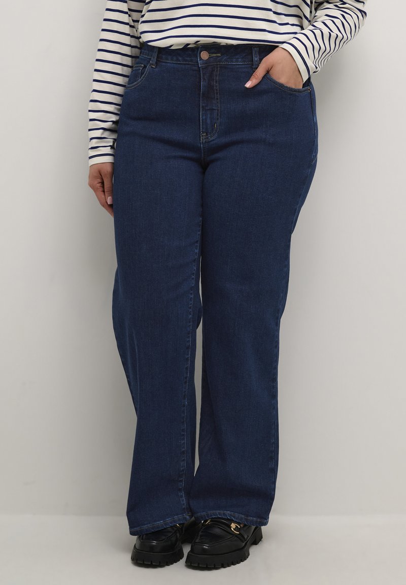 Jeans anchos de color azul oscuro con una cintura alta, bolsillos delanteros y costuras en contraste; combinados con una camisa de rayas de manga larga y zapatos negros.