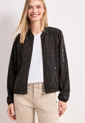 Smilende kvinde iført en sort og mørkebrun bomberjakke med leopardprint over en hvid skjorte og beige bukser, stående mod en ensfarvet lys baggrund.