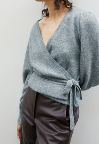 Ljusgrå, omslutande tröja i mjukt, fluffigt material med djup V-ringning och knytmidja. Kombineras med mörkbruna, släta läderbyxor.