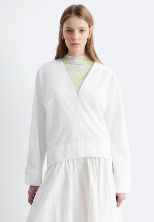 Mladá žena s dlhými vlasmi, ktorá má na sebe bielu bundu s výstrihom do V cez vzorovaný top s vysokým golierom a bielu sukňu na jednofarebnom pozadí.