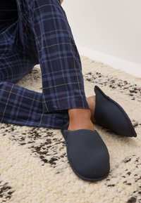 Ember kék kockás pizsamanadrágot és sötétszürke, texturált papucsot visel, krém és barna mintás szőnyegen ül.