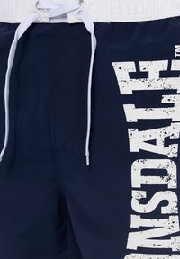 Navy swim shorts with white elastic waistband, drawstring, and large distressed text graphic on the right leg. Smooth fabric, knee-length fit.