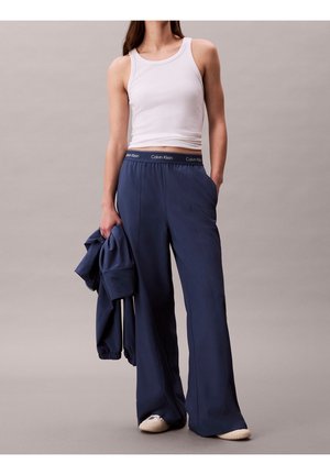 Mujer vistiendo una camiseta blanca sin mangas y pantalones sueltos azul marino Calvin Klein, sosteniendo una chaqueta azul marino a juego contra un fondo liso.