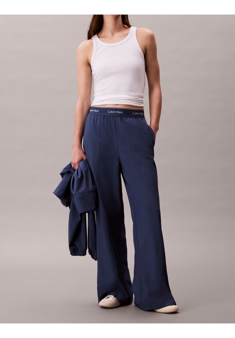 Femme portant un haut blanc sans manches et un pantalon ample bleu marine Calvin Klein, tenant une veste assortie bleu marine sur un fond uni.