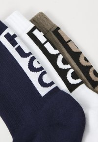 Trois paires de chaussettes côtelées en bleu marine, blanc et marron, avec un logo tricoté dans le revers en couleurs contrastées.