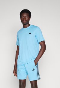 Svijetloplava sportska majica s crnim Adidas logotipom, uparen s odgovarajućim kratkim hlačama s crnim bočnim prugama. Glatka, prozračna tkanina.