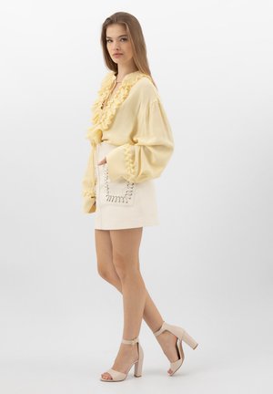 Vrouw in een lichtgele blouse met ruches en een crèmekleurige mini-rok met gestikte zakdetails, draagt beige enkellaarsjes met blokhak, staand in zijaanzicht.