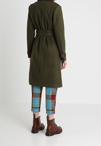 Olivengrüner, taillierter Mantel mit strukturiertem Finish, kombiniert mit karierter, verkürzter Hose in Blau, Rot und Gelb sowie braunen Schnürstiefeln.