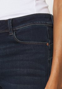 Jeans de denim azul oscuro con corte slim, costuras visibles, un bolsillo frontal y un detalle de remache de cobre en la cintura.