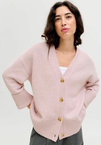Cárdigan de punto en un suave tono rosa claro con escote en forma de V, botones de madera, hombros caídos y puños acanalados. Presenta un ajuste ligeramente oversize y aberturas laterales.