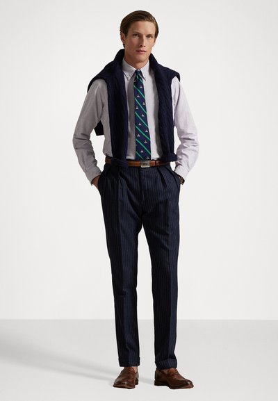Pantalon bleu marine à rayures, chemise à rayures claires, cravate verte avec des accents bleus, pull en maille bleu marine drapé sur les épaules, chaussures marron.