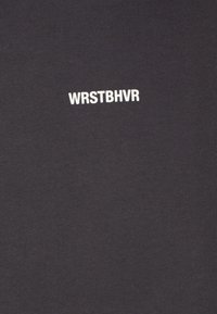 Camiseta negra de canalé con el texto blanco "WRSTBHVR" en una fuente moderna en el pecho, confeccionada con una suave y elástica mezcla de algodón.