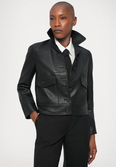 Zwarte leren cropped jas met knoopsluiting aan de voorkant, twee voorzakken, staande kraag en slim-fit silhouet. Gecombineerd met een witte blouse en stropdas.