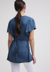 Niebieska tunika z krótkimi rękawami, z falbanami i wiązanym detalem w talii, wykonana z miękkiego materiału, noszona na białych spodniach.