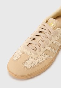 Beżowe buty sportowe z plecioną cholewką, gładkimi akcentami z gumy i wzorem w trzy paski. Posiadają płaskie sznurowadła i metkę z logo.
