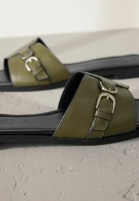 Sandales à enfiler en cuir vert avec une large lanière ornée d'une boucle argentée, semelle plate noire et finition lisse texturée.