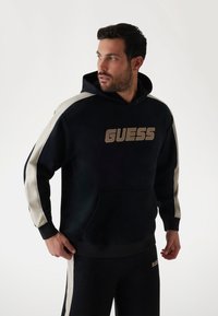 Čierna mikina s kapucňou s kontrastnými krémovými bočnými pruhmi a predným vreckom. Obsahuje veľké logo "GUESS" v béžovej farbe na hrudi.