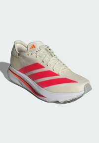 Béžová sportovní obuv se třemi růžovými pruhy, bílou podrážkou a oranžovými akcenty. Má texturovaný svršek a design s šněrováním.