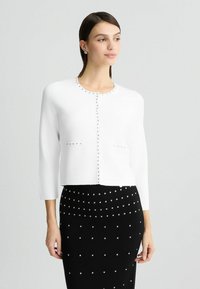 Cardigan bianco con scollatura rotonda, maniche a tre quarti e borchie argentate lungo il bottoniere; abbinato a una gonna nera aderente con dettagli in perle.