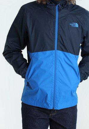 Mężczyzna ubrany w czarno-niebieską kurtkę North Face na suwak, z rękami w kieszeniach, w ciemnych dżinsach.